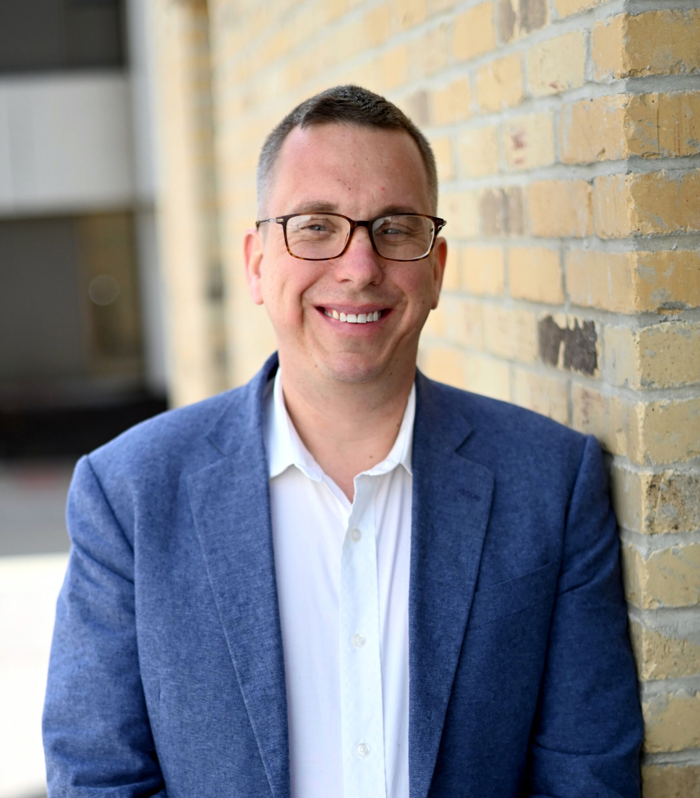 Portrait of a man wearing glasses and a blue blazer, smiling while leaning against a beige brick wall.