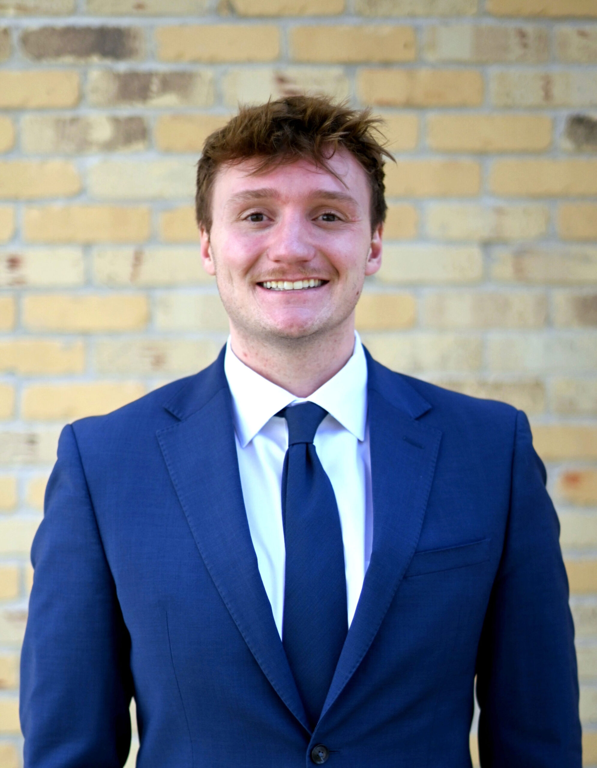 Tyler Timm Profile Picture - Kittelsen Law | Law Firm in Monroe providing legal services Smiling man in a blue suit and tie posing against a yellow brick wall.