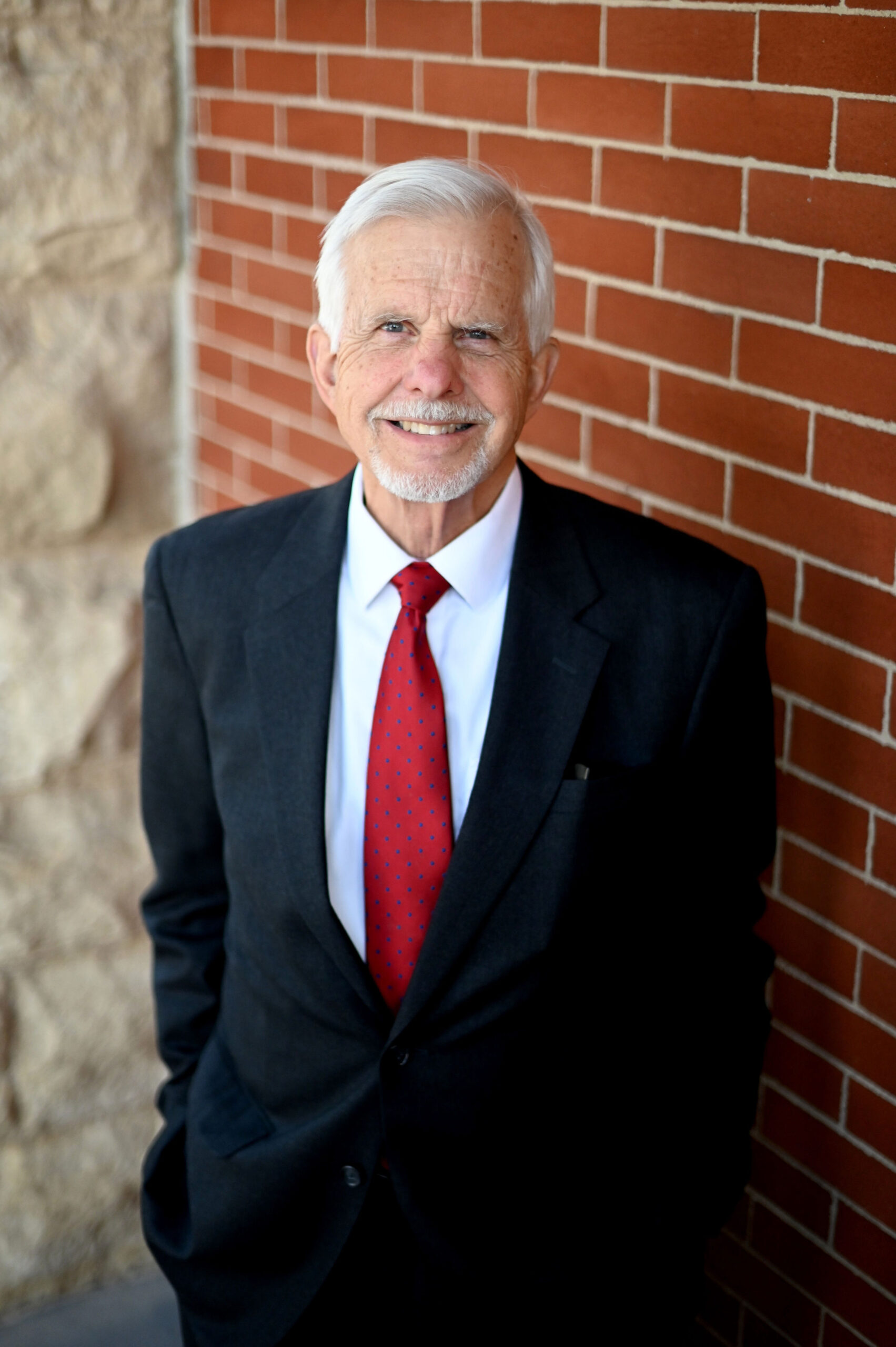 Smiling older man in a dark suit and red tie stands against a brick wall.