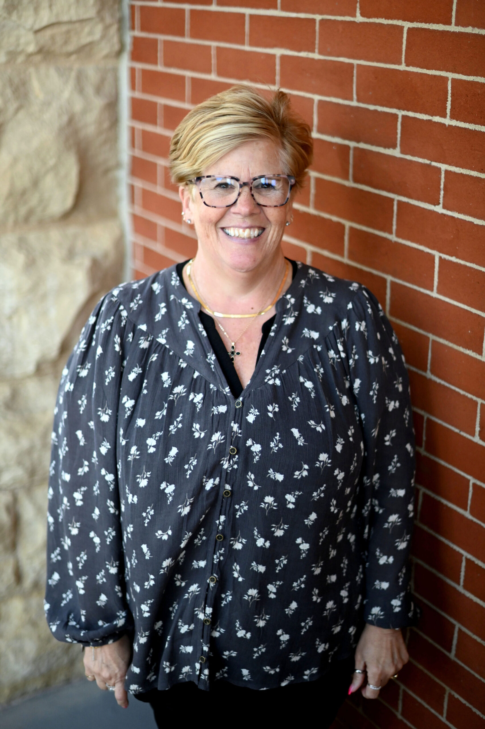Smiling woman with short blonde hair and glasses stands against a red brick wall, wearing a dark floral blouse and layered necklaces.