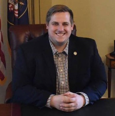 Taylor Call - Kittelsen Law | Law Firm in Monroe providing legal services Smiling man in a dark blazer sits at a desk in an office, hands folded, with a flag in the background.